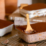 Chocolate tiramisu desserts in rectangular metal molds, with one in the foreground on a wire cooling rack and a wooden spoon resting in it. The scene includes chocolate pieces on a wooden surface, with blurred ceramic containers and more desserts in the background.