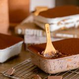 Chocolate tiramisu desserts in rectangular metal molds, with one in the foreground on a wire cooling rack and a wooden spoon resting in it. The scene includes chocolate pieces on a wooden surface, with blurred ceramic containers and more desserts in the background.
