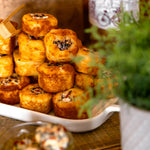 Stacked golden-brown mini pastries or tarts on a white serving tray, with a glass bowl of similar items in the foreground and a potted green plant on a rustic wooden table.