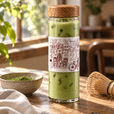 A clear glass bottle with a wooden lid and a white illustrated label, filled with green iced matcha drink, set on a wooden table in a warm, sunlit room with a bowl of matcha powder and a bamboo whisk nearby.